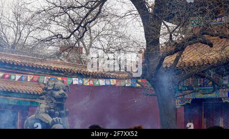 Eine Löwensteinstatue vor dem alten China Palast mit roter Mauer und umgebenen Ästen, Rauch fließt in die Luft Stockfoto