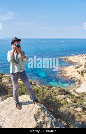 Ein junger Tourist macht ein Foto von der Spitze eines Hügels, wo man das Meer mit schönen Farben sehen kann, trägt er einen sehr coolen Look Stockfoto