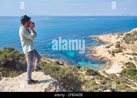 Ein romantischer junger Mann fotografiert mit seiner Filmkamera In einer Bucht des Mittelmeers in einem touristischen Bereich, wo das Meer hat schöne Farben Stockfoto