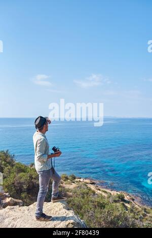 Ein Reisender genießt die Aussicht von der Spitze eines Hügels, wo er das Meer sehen kann, er trägt einen Hut, ein Hemd und Jeans, es ist tagsüber und der Himmel ist clea Stockfoto