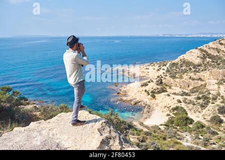 Ein junger Tourist macht ein Foto von der Spitze eines Hügels, wo man das Meer mit schönen Farben sehen kann, trägt er einen sehr coolen Look Stockfoto