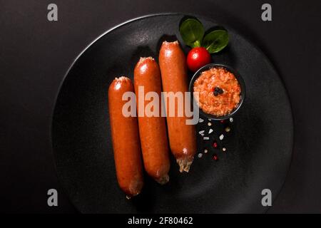Gegrillte Würstchen mit Salat und Sauce auf einem schwarzen Teller Mit schwarzem Hintergrund Stockfoto