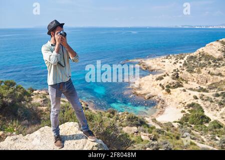 Ein junger Tourist macht ein Foto von der Spitze eines Hügels, wo man das Meer mit schönen Farben sehen kann, trägt er einen sehr coolen Look Stockfoto