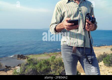 Ein nicht erkennbarer Reisender verwendet EIN SMARTPHONE, WÄHREND er ein hält Vintage-Kamera mit dem Meer im Hintergrund Stockfoto