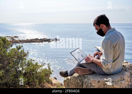 Ein junger Mann arbeitet ferngesteuert mit seinem Laptop auf einem Felsen auf einem Hügel mit dem Meer im Hintergrund Stockfoto