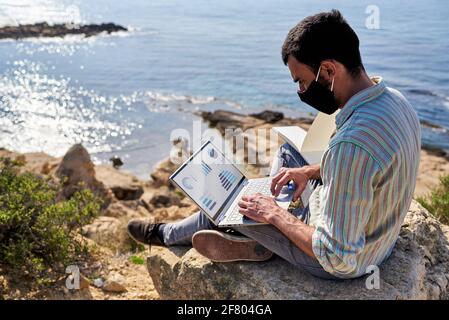 Ein junger Mann arbeitet ferngesteuert mit seinem Laptop auf einem Felsen auf einem Hügel mit dem Meer im Hintergrund Stockfoto