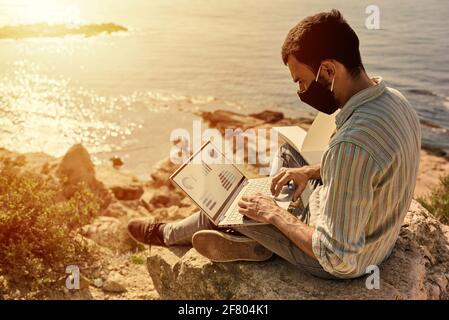 Ein junger Mann arbeitet ferngesteuert mit seinem Laptop auf einem Felsen auf einem Hügel mit dem Meer im Hintergrund Und der Sonnenuntergang über dem Meer Stockfoto