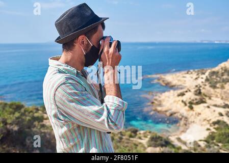 Ein junger Tourist macht ein Foto von der Spitze eines Hügels, wo man das Meer mit schönen Farben sehen kann, trägt er einen sehr coolen Look Stockfoto