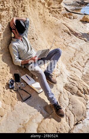 Ein junger Mann entspannt sich, indem er seinen Hut vor sein Gesicht legt, während er ein Nickerchen macht, er hält seinen Laptop und an seiner Seite hat er eine Vintage-Kamera und eine Stockfoto