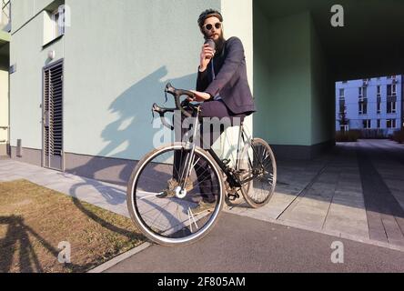 Starten Sie mit frischem Kaffee in den neuen Tag. Millennial Geschäftsmann hält eine Tasse Kaffee und lehnt sich an sein Fahrrad und schaut auf den freien Raum. Stockfoto