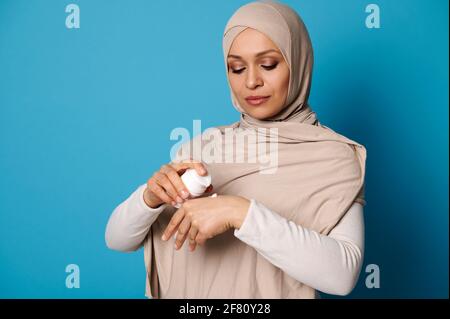 Porträt einer schönen arabischen muslimischen Frau in Hijab, die Creme auf der Hand quetscht, isoliert auf blauem Hintergrund mit Platz für Text Stockfoto