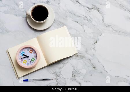 Runder rosafarbener Wecker, quadratisches Notizbuch und Stift auf Marmortischoberfläche. Tasse Kaffee. Speicherplatz kopieren Stockfoto