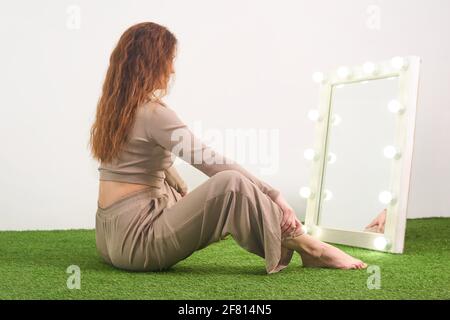 Frau, die ihre Spiegelung in einem beleuchteten Spiegel anschaut, um sich zu kleiden Das Zimmer befindet sich auf dem Boden im Studio Stockfoto