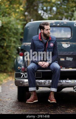 Berded junger Mann in Jeans sitzt auf seinem Pickup chevrolet Oldtimer draußen .junger Mann sitzt auf der Rückseite eines alten Chevrolet Pick-up-Truck. Stockfoto