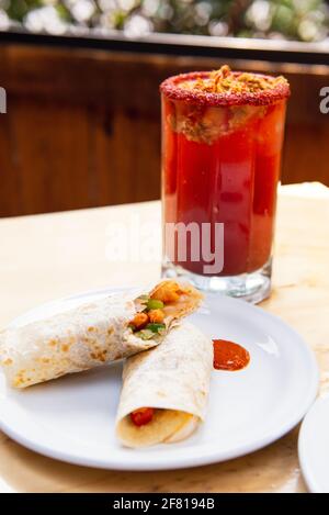 Vertikale Aufnahme von Burrito mit Clamato auf einem weißen Teller Und Bloody mary in einem durchsichtigen Glas Stockfoto