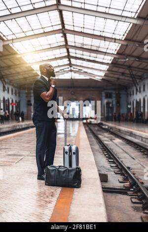 Vertikale Aufnahme eines eleganten afroamerikanischen Unternehmers, der seine Einstellung anpasst Krawatte, während mit Taschen auf der Bahnsteig von stehen Ein Bahnhofsdepot Stockfoto