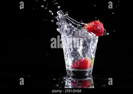 studiofoto von Erdbeeren und Eis, die vor schwarzem Hintergrund in ein Wasserglas gefallen sind. Stockfoto