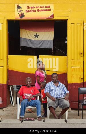 2 Männer sitzen vor einer wunderbar bunten Bar mit gelben Wänden und einer ghanaischen Flagge, die am Eingang in Cape Coast, Ghana, hängt Stockfoto