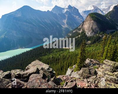 Hoher Berggipfel neben einem türkisfarbenen See in der rockies mit einem Pinienwald und Felsen in der Im Sommer im Vordergrund Stockfoto