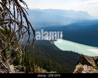 Großer türkisfarbener See unten in den Bergen mit Nebel In der Ferne im Sommer und Äste im Vordergrund Stockfoto