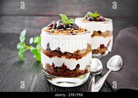 Dessert Schwarzwald von Kirschen, Schokoladenkeks und weichen Quark mit Sahne in zwei Gläsern, Serviette, Minze und Löffel auf Holzbrett Backg Stockfoto