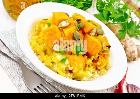 Hirsebrei mit würziger Kürbissauce und Samen auf einem Teller auf Serviette, scharfer Pfeffer, Ingwerwurzel, Knoblauch und Petersilie auf dem Hintergrund von hellem Holz Stockfoto