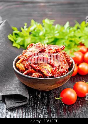 Sonnengetrocknete Tomaten in Öl mit Thymian und Basilikum in einer Schüssel, Serviette, frische kleine Tomaten und Petersilie auf schwarzem Holzbrett Hintergrund Stockfoto