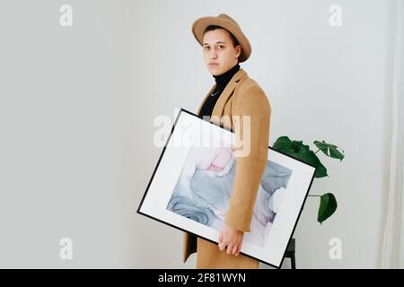 Stilvoller junger Mann in beigefarbenem Anzug und klassischem Hut, der mit abstrakter Malerei unter seinem Arm posiert. Er steht seitlich und schaut auf die Kamera. Indoo Stockfoto