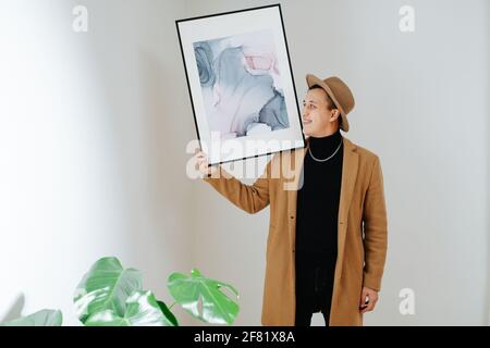 Lächelnder Geschäftsmann in einem beigen Anzug und einem klassischen Hut, der mit abstrakter Malerei auf seiner Schulter posiert. Er malt mit Blick auf die Kamera und schaut sie an. Zoll Stockfoto