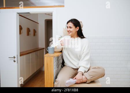 Hübsche junge Frau, die mit dem Bein nach oben auf einem hohen Beistelltisch sitzt und aus einem Becher trinkt. Sie lächelt und sieht jemanden an. Lässig gekleidet. Stockfoto