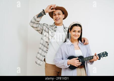 Erhabene junge Paare, die nahe beieinander stehen, ine hinter dem anderen. Frau spielt kleine Gitarre. Vor der weißen Wand. Er trägt einen Strohhut Stockfoto