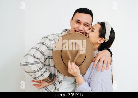 Entzückendes junges Paar, das dicht beieinander steht und den unteren Teil des Gesichts mit einem Hut bedeckt. Vor der weißen Wand. Mann umarmt Frau, drücken sie Stockfoto