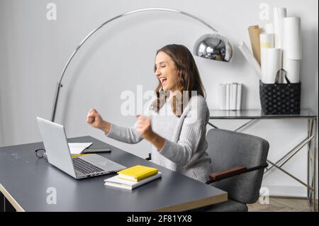 Aufgeregt junge Frau in Freizeitkleidung Schrei ja sitzen vor einem Laptop im modernen Büro, weibliche Mitarbeiter feiern besten Deal, Stellenangebot, gewinnen in Online-Lotterie, die Arme glücklich zu heben Stockfoto