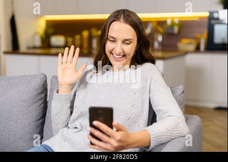 Videoverbindung über die Distanz. Positive junge Frau, die das Smartphone für Videoanruf nutzt. Lächelnde Frau hat ein angenehmes Online-Meeting, sitzt auf der Couch zu Hause, genießt Online-Chats mit Freunden Stockfoto