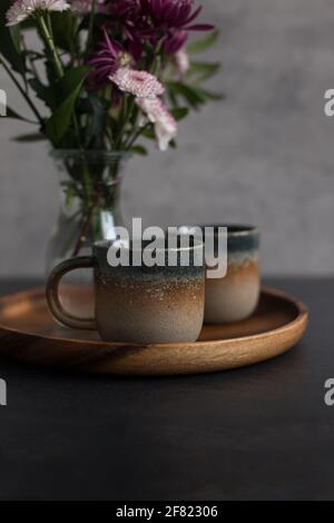 Zwei elegante, rustikale Kaffeetassen auf einem Holztablett neben einem verschwommenen Bouquet von Dahlien. Stockfoto