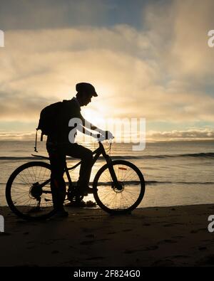 Silhouettenbild eines Radfahrers, der sich bei Sonnenaufgang auf den Strand vorbereitet. Vertikales Format. Stockfoto