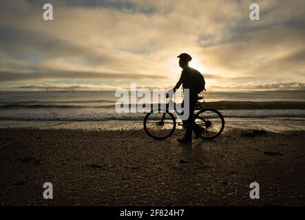 Silhouette Bild eines Radfahrers zu Fuß und schieben das Fahrrad Am Strand bei Sonnenaufgang Stockfoto