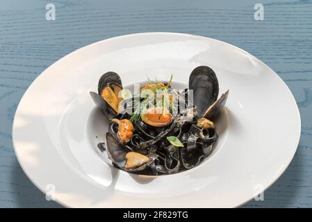 Schwarze Tintenfisch-Tinte mit Muscheln - italienische Tagliatelle al nero di seppia - in einem weißen Gericht auf blau Holztisch Stockfoto