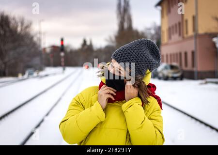 Frau mit Gesichtsmaske wartet im Winter auf den Zug am Bahnhof. Reisen während des Coronavirus, einer Covid-19-Pandemie. Junge Frau mit gelbem Mantel Stockfoto