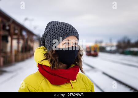 Junge Frau mit Gesichtsmaske und steht im Winter am Bahnhof. Reisen während des Coronavirus, einer Covid-19-Pandemie. Stockfoto