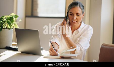 Eine reife Frau am Schreibtisch macht sich Notizen, während sie mit dem Handy spricht. Geschäftsmann spricht mit dem Kunden auf dem Smartphone und schreibt in der Molkerei. Stockfoto