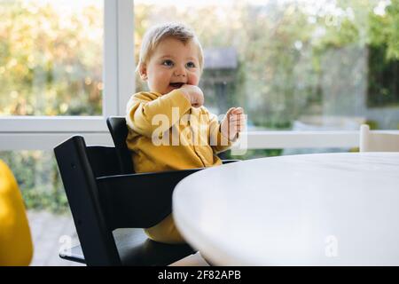 Netter kleiner Junge, der auf einem Stuhl am Tisch sitzt. Kind wartet auf Essen. Niedliches Baby, das auf einem Hochstuhl sitzt und darauf wartet, zu essen. Stockfoto