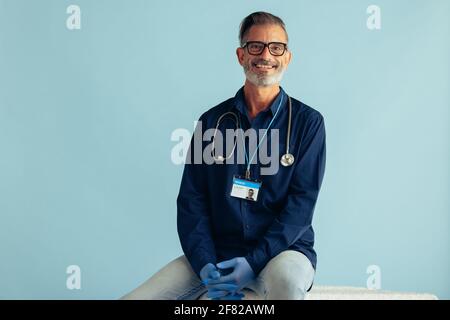 Porträt eines erfahrenen reiferen Arztes, der vor blauem Hintergrund sitzt. Allgemeinpraktizierender Arzt mit Stethoskop, der die Kamera ansah und lächelte. Stockfoto
