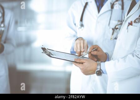 Unbekannte Ärzte, zwei Männer und eine Frau, diskutieren medizinische Untersuchungsresoults, während sie im Krankenhaus-Büro stehen. Ärzte, die Klemmbretter zum Auffüllen verwenden Stockfoto