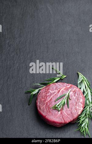 Frisches rohes Rindersteak mit Rosmarinstift auf schwarzem Stein Hintergrund Stockfoto