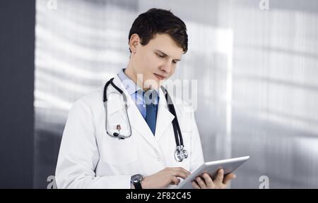 Ein junger, freundlicher Arzt überprüft einige Informationen auf seinem Computer-Tablet. Porträt eines Facharztes bei der Arbeit in einer Klinik. Medizinkonzept Stockfoto