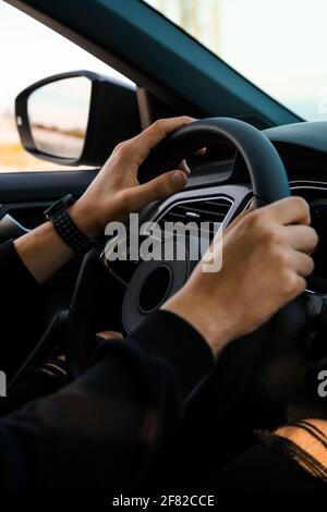 Nahaufnahme der Hände, die den Lenker eines Autos halten, mit einer intelligenten Uhr am Handgelenk. In dunkler, moderner Kleidung. Stockfoto