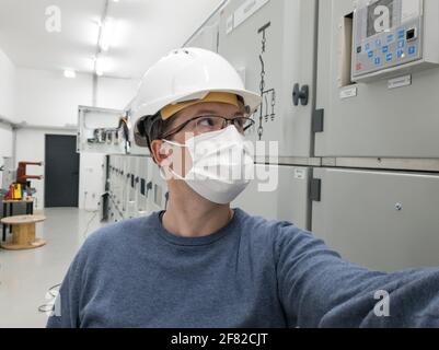 Junger Ingenieur, der in einer Elektrounterstation arbeitet und eine Gesichtsmaske trägt. Stockfoto