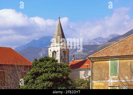 Mediterranes Dorf: Rote Ziegeldächer und Glockenturm gegen Himmel und Berge. Montenegro, Tivat, Dorf Donja Lastva, katholische Kirche St. Ro Stockfoto