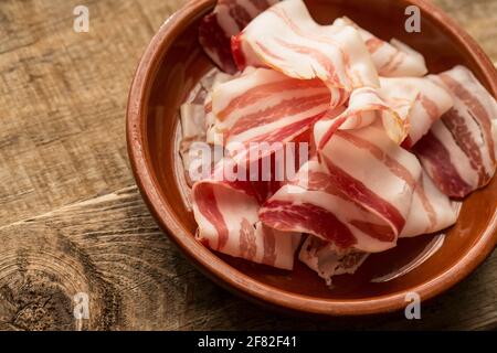 Dünne Scheiben luftgetrockneter roher italienischer Pancetta, die aus einem Supermarkt gekauft wurden und aus einer Plastikverpackung entfernt wurden. In einer Schüssel auf einem Holz dargestellt Stockfoto